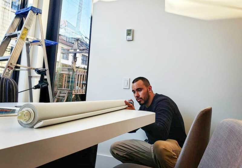 AP shades employee in casual attire kneels by a desk, examining a roll of shades material in a modern office setting with construction visible through the window.